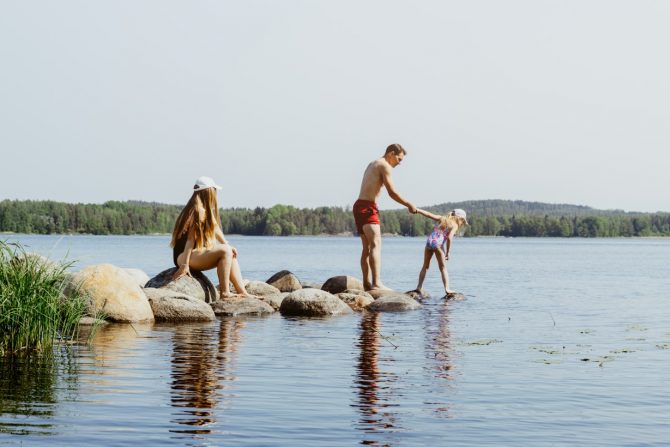 Nainen, mies ja lapsi järven rannalla kesällä.