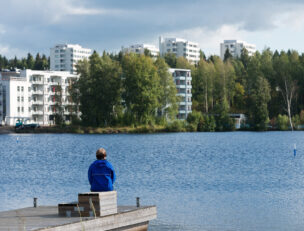 Henkilö järven rannalla, taustalla kerrostaloja.