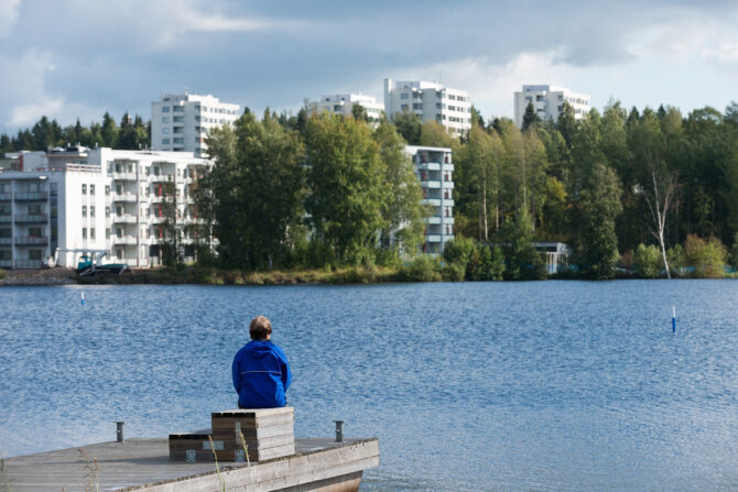 Henkilö järven rannalla, taustalla kerrostaloja.