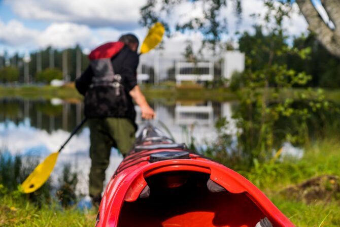 Henkilö vetämässä kanoottia lammelle, taustalla Mikaelin rakennus.