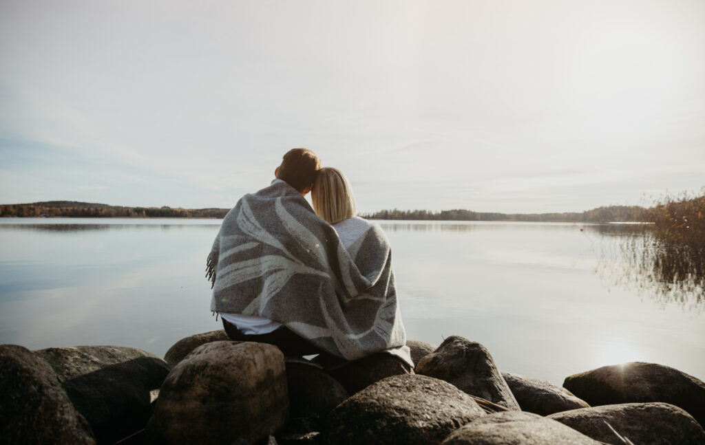 Mies ja nainen istuvat rantakivetyksellä selät kameraan päin. Taustalla järvimaisema.